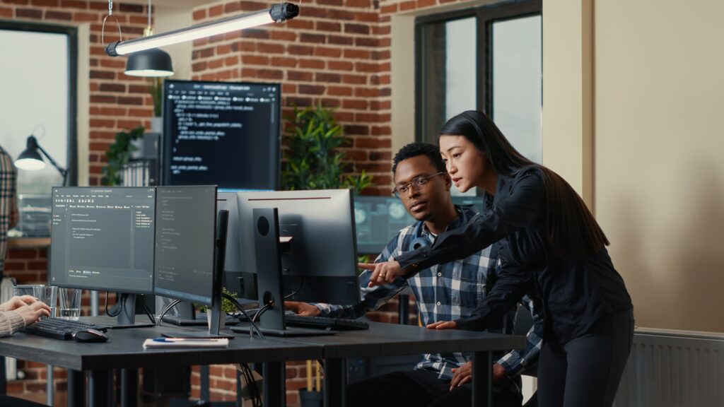 Two software developers holding laptop with coding interface walking towards desk and sitting down